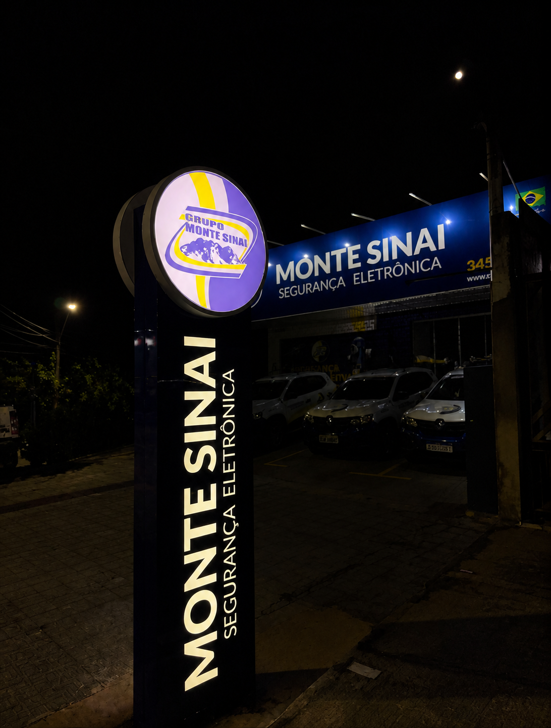 Totem completo do Grupo Monte Sinai iluminado à noite, com fachada e viaturas ao fundo